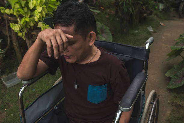 A disabled and poor asian man in a wheelchair covers his forehead with his hand. Stressed and worried. Rural setting. A disabled and poor asian man in a wheelchair covers his forehead with his hand. Stressed and worried. Rural setting. polio virus photos stock pictures, royalty-free photos & images