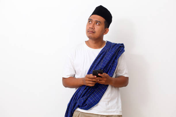 istock Thoughtful asian man while standing, holding a cellular phone and looking at blank space. Isolated on white background 1409010885