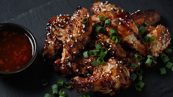 Grilled chicken wings with sauce garnished with green onions on slate board over black stone background.