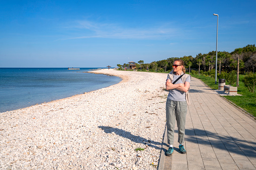 Senior woman enjoying nature by standing on pathway by the beach in Peroj near Pula in Istria. It is morning, relaxing time, good for exercising and meditation. In background is blue sky and long, long beach, Croatia, Europe.