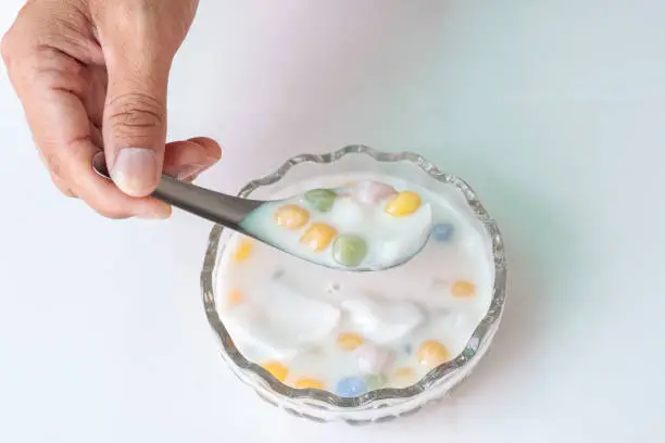 Photo of Hand holding spoon to scooping rice balls with young coconut in sweet coconut milk in a cup isolated on white  tile background Thai people call Bua Loi.