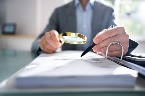 Close-up Of A Businessperson's Hand Looking At Receipts Through Magnifying Glass At Workplace