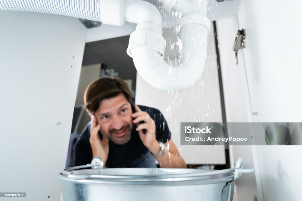 Worried Man Calling Plumber Worried Man Calling Plumber While Watching Water Leaking From Sink Plumber Stock Photo