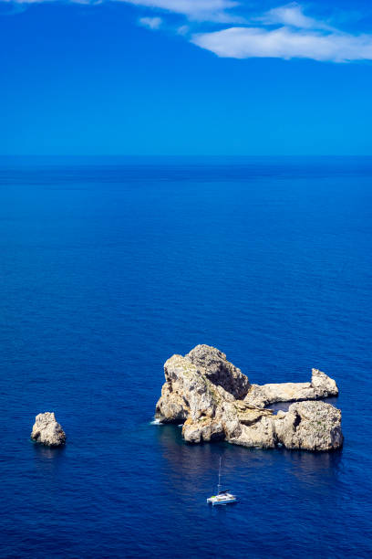 View of the islets of Ses Margalides, off the western coast of Ibiza. The bright light of a summer noon, deep blue waters as far as the eye can see, picturesque clouds, a moored sailing boat. Developed from RAW.
