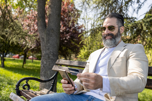 Cheerful man smiling when sitting on park bench and using credit card and smart phone