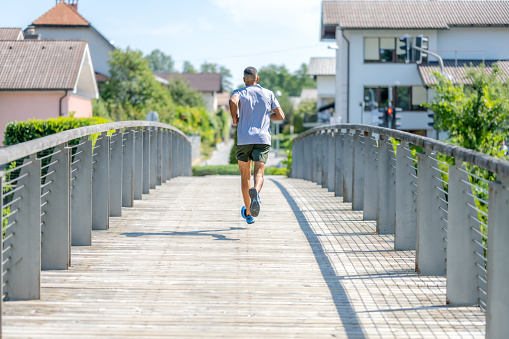 African American sporty man jogging outdoors, back view. Sportsman running across bridge on summer sunny day, wide shot.