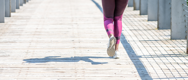 Fit woman in sportswear running back view, close up on legs and sneakers. Girl outdoor workout on sunny day. Sportswoman training