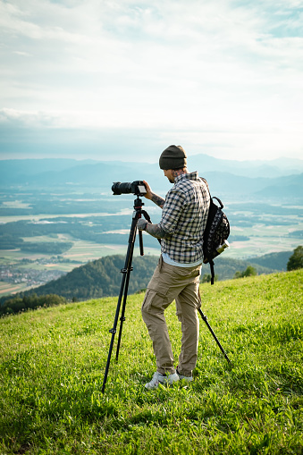 Landscape photographer working with tripod in beautiful green nature, standing with backpack on top of the hill.