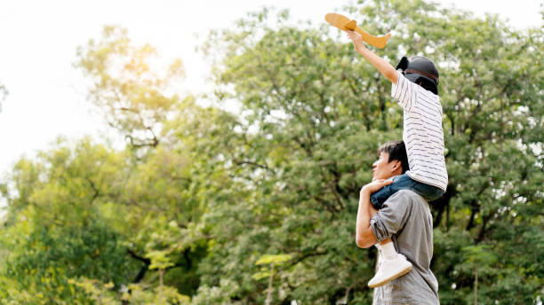 Lifestyle Asian family Father and son sat on his shoulders and run in park. Paper plane as a toy in hand of kid And there are many big trees in garden Is natural in the morning of summer stock photo