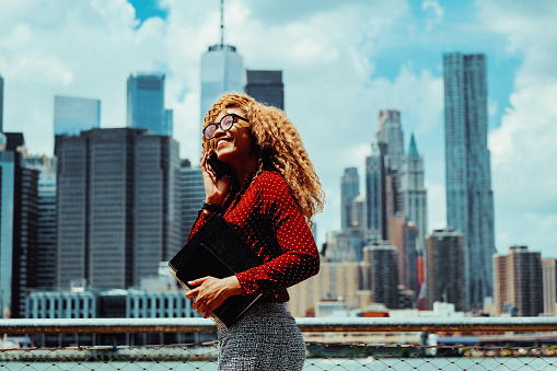 Portrait smiling young adult entrepreneur millennial woman with eyeglasses and afro hair talking on a phone call outdoors with Manhattan New York City skyline skyscraper behind Hudson river