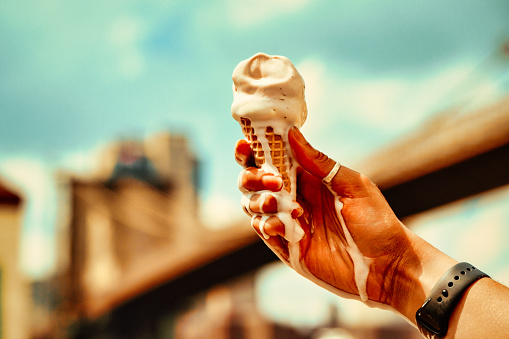 Melting vanilla ice cream cone on hand against blue sky and Brooklyn bridge in the background shot