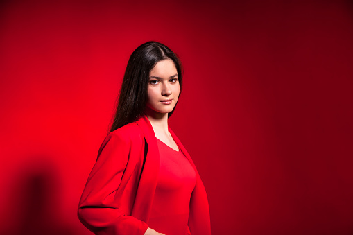 Positive teen girl in stylish red costume over red background looking at camera. Young beautiful cute brunette lady standing and posing. Emotion concept. Copy space