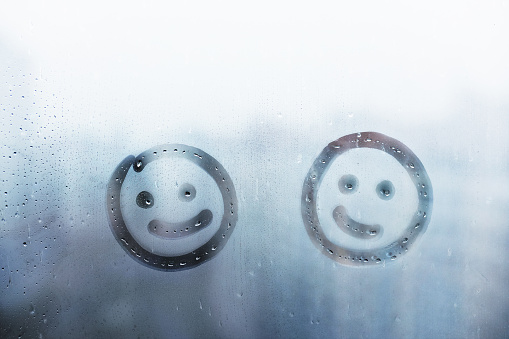Two happy smile doodle face painted on blue window flooded with raindrops