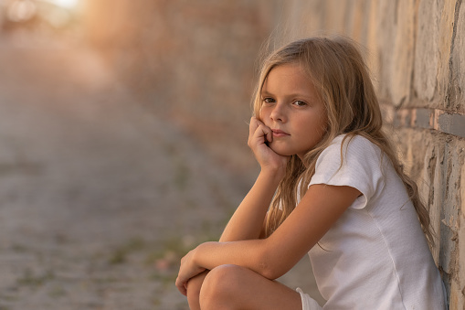 Close-up portrait of a young girl with a sad look. The child pressed his back against a stone wall and looks somewhere into the distance.