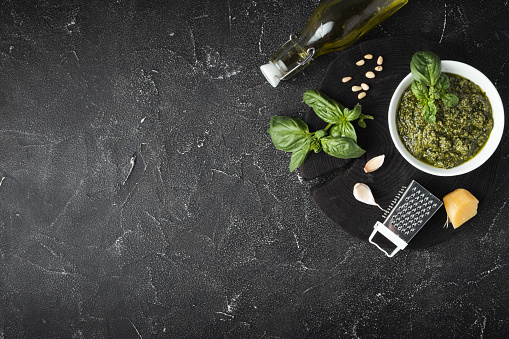 Ingredients for pesto sauce in the white bowl. Green basil leaves, Parmesan cheese and pine nuts on the black background. Flat lay. Overhead view