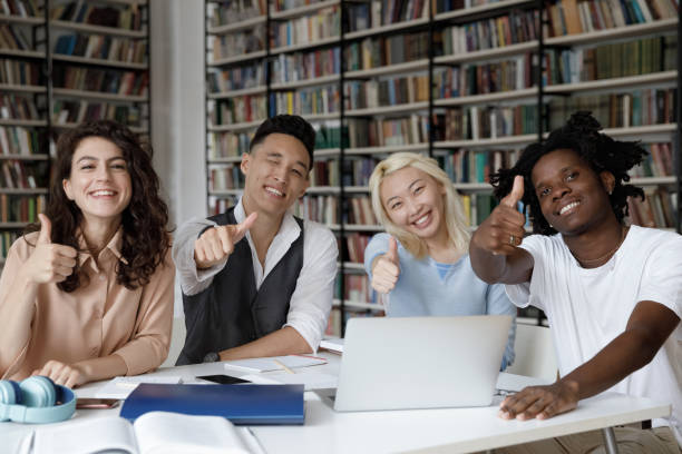 многоэтнические студенты, показывая большие пальцы вверх, сидя�т за столом в библиотеке - students exam results стоковые фото и изображения