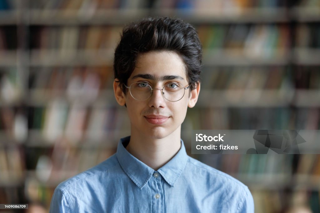 Head shot portrait student guy in glasses posing in library Head shot schoolboy guy posing in campus library on bookshelves background. 17s pupil in eyeglasses look at cam. Excellent student portrait, skill and knowledge, education, generation Z person concept Judaism Stock Photo