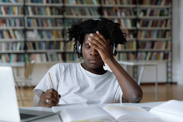 African student feels tired due long preparations for college exam African student feels tired due to long preparations for college exam, deadline task, suffers from headache sit at table with laptop in library. Information overload during admission, stress concept information overload photos stock pictures, royalty-free photos & images