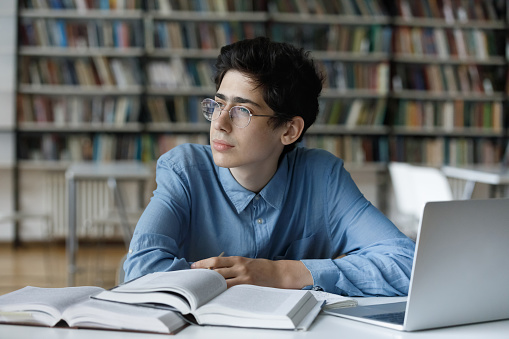 Pensive student guy in glasses staring aside, sit at table in library, distracted from studies, preparation for university admission or college exams looks thoughtful. Education, work on essay concept