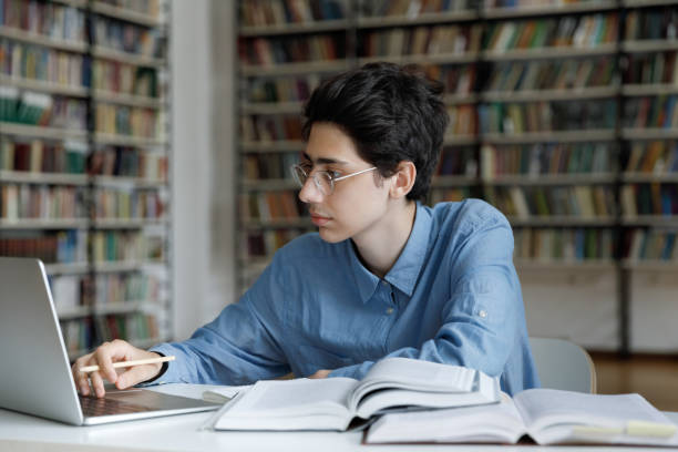 Student guy studying, sits at table with laptop in library Student guy works on summary or essay, busy in exams preparations sit at table with books use laptop studying in university library. Young gen use modern tech to gain new knowledge, education concept book summary apps stock pictures, royalty-free photos & images