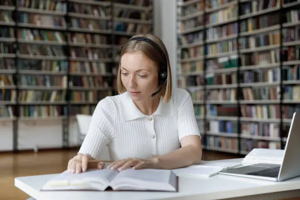 Pretty student teen girl in headset studying sit at table with books and laptop in library. Preparation for exams use modern tech, audio course to improve foreign language knowledge. Education concept