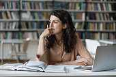 Thoughtful girl studying in library seated at table with textbooks