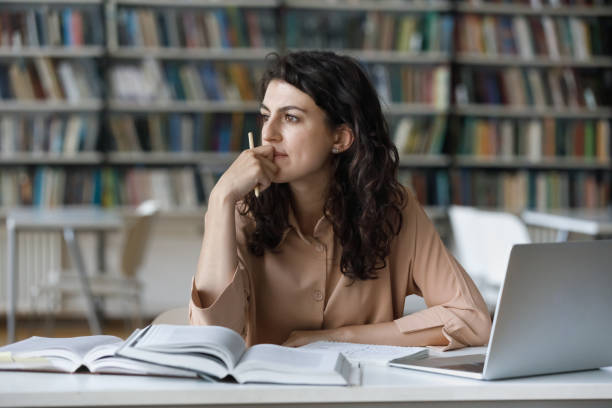 Thoughtful girl studying in library seated at table with textbooks financial markets Young student girl sit at table with textbooks and laptop staring aside, studying alone in library, looks pensive and thoughtful search solution, prepare for exam, makes task feels confused or puzzled Decision Making stock pictures, royalty-free photos & images