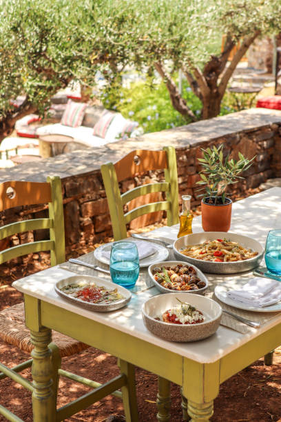 греческий обеденный стол с едой и тарелками под оливковыми деревьями с критскими деликатесами - средиземная пища стоковые фото и изображения