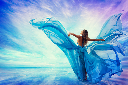 Woman in Blue Dress Flying on Wind looking at Sky and Sea. Beautiful Model Arms outstretched enjoying Freedom at Beach Summer Resort Rear view. Artistic Women Silhouette in Fantasy Gown as Butterfly Wings. Art Female Portrait looking away at Sunrise Landscape