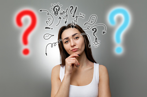 The concept of searching for ideas and information. Portrait of a pensive young Caucasian woman with her hand on her chin. Gray background and light with drawn question marks and symbols