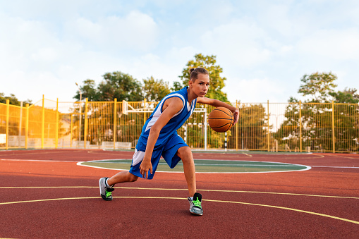 Basketball. A teenage boy in blue sportswear plays a basketball. In the background is a sports field. Copy space. Concept of sports games.