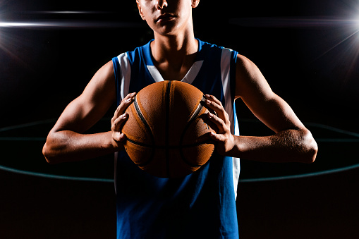 Basketball. Close up portrait of a teenage boy in blue sportswear confidently holds a basketball with muscular hands. Black background. Concept of sports games.