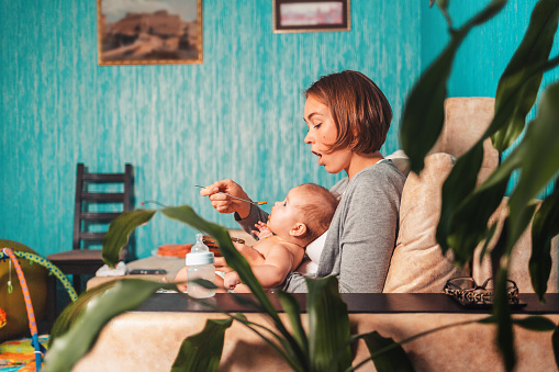 Young mother is sitting on the couch with the baby, and feeds it with a spoon. There is a feeding bottle nearby. Side view. The concept of feeding and weaning baby from the breast.