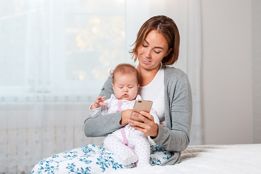 A mother holds a sleeping child in her arms and shows him something on her smartphone. Close-up portrait. Home interior.