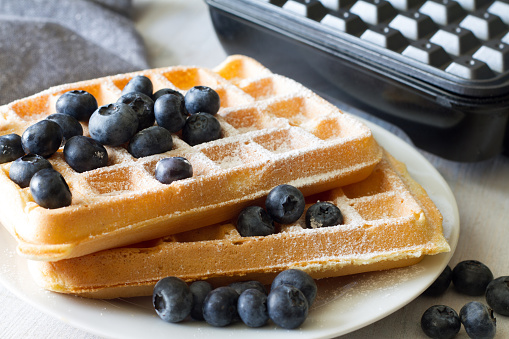 Delicious waffles with blueberries and powdered sugar, waffle maker in background concept