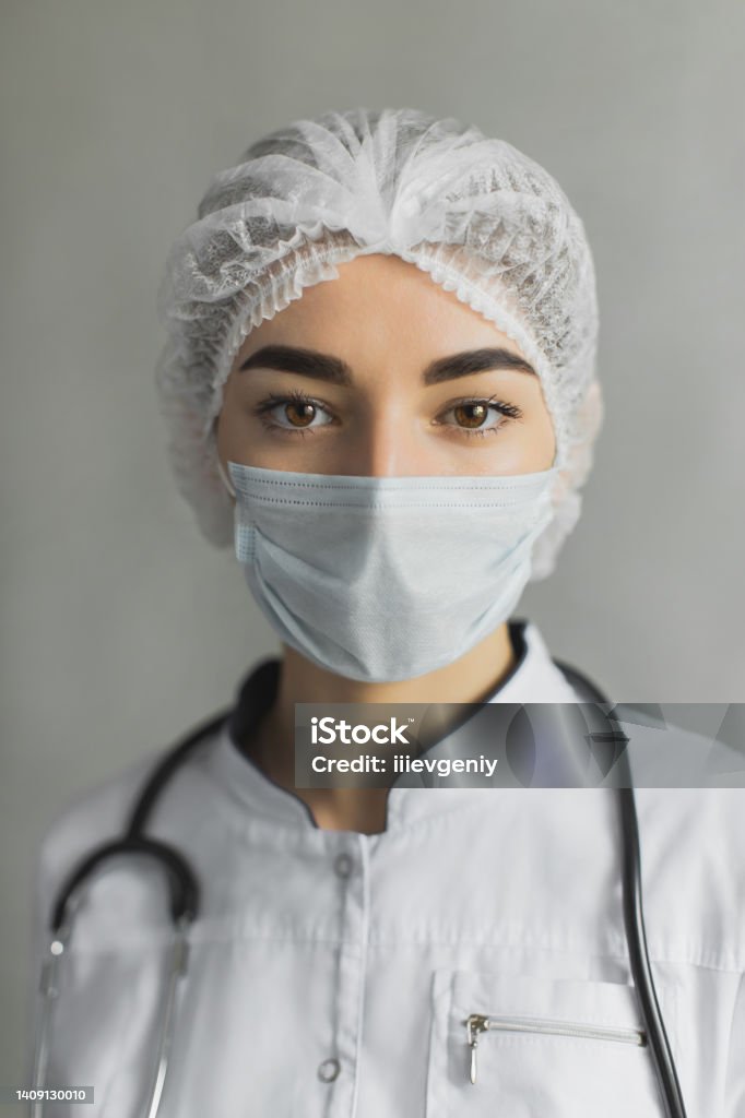Young doctor portrait in white coat and protective mask on gray background. Medical specialist in professional uniform. Beautiful brunette girl in protective gear. Confident nurse Cut Out Stock Photo