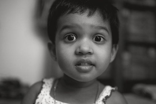 Monochromatic portrait of a little Bengali (Indian ethnicity) girl with expressive face, looking at camera. Photo taken inside her room.