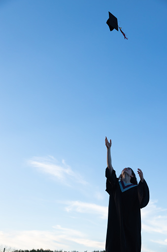 Teen girl with curly dark hair wearing a graduation cap and gown outdoors throwing cap into the air against a bright blue sky with copy space