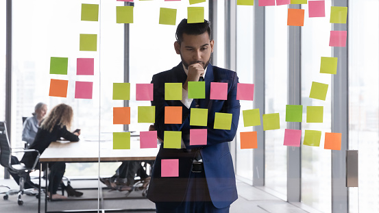 Young pensive Indian businessman in suit stand near glass wall staring at attached adhesive colored memory sticky post-it reminders notes with visual tasks, business ideas, new start-up looks focused