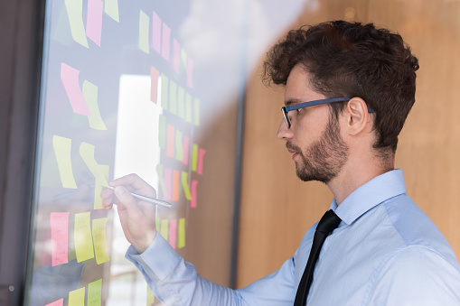 Profile side view focused employee businessman holds pen write down necessary information on multicolored sticky notes papers attached on glass wall, recording every day tasks, create strategy concept