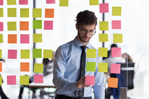 Serious millennial businesswoman stand in office writes on sticky notes on glass wall prepare boardroom for negotiations or briefing. Start up ideas, develop strategy, make schedule or agenda concept