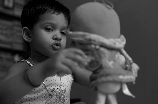 Monochromatic portrait of a little Bengali (Indian ethnicity) girl with expressive face, playing with a soft toy. Photo taken inside her room.