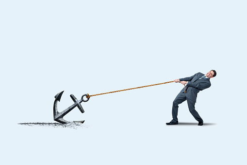 A businessman is burdened with the prospect of pulling on a rope that is tethered to an anchor as he attempts to drag it on his way to his destination.
