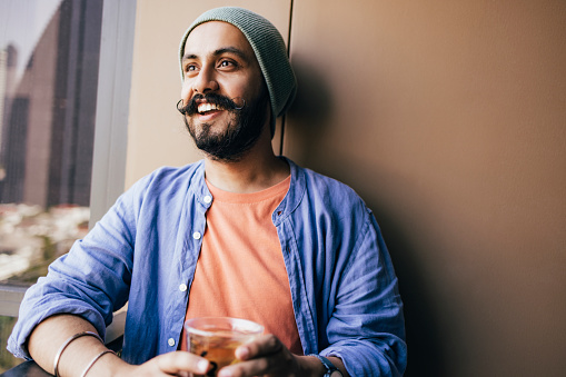 A smiling Indian male with a beanie looking away while drinking tea on the balcony.