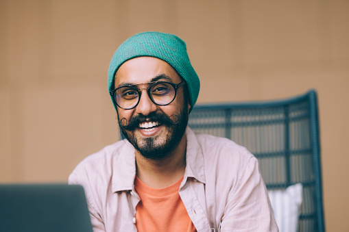 A portrait of a happy Indian entrepreneur with glasses and a beanie working in his office.