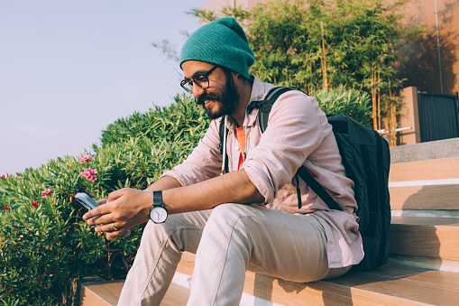 A handsome bearded Indian hipster with glasses and a green beanie smiling while texting  on his smartphone.