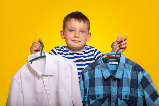 istock Cute little kid boy trying to decide between two shirt on yellow background. A boy holds two hangers with shirt in his hands 1409150953