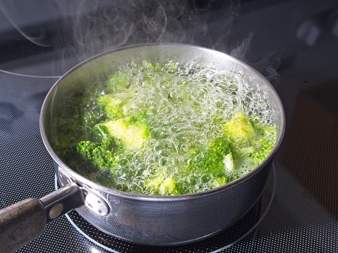 Boiling broccoli in a cook pan on a stove. Domestic life of cooking nutritious vegetables for dinner, with steam rising from pot.