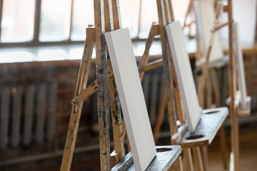 Wooden easels with white blank canvas in row inside cozy loft room, no people. Academy of art education, hobby, training in group class for creative skill development, artistic school tuition concept