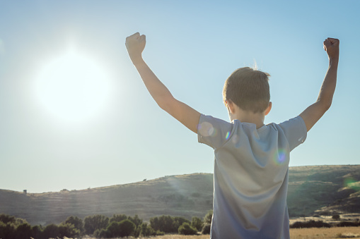 A young boy is raising both fists in victory with a bright sun and blue sky in the background. Image taken on Lemnos Greece.
